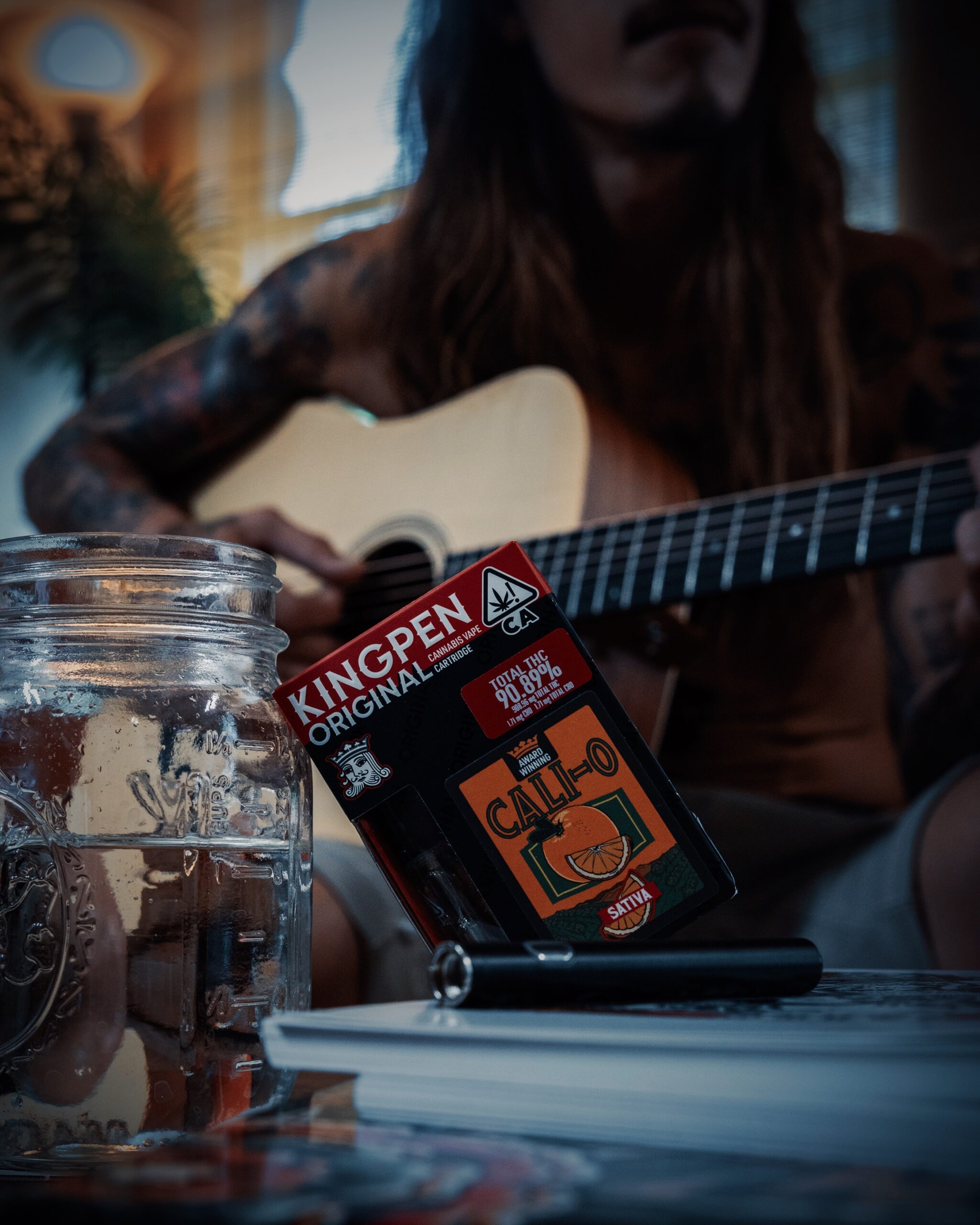 Kingpen vape cartridge in award winning Cali O sativa sits next to a glass of water in foreground, tattooed man plays acoustic guitar in background