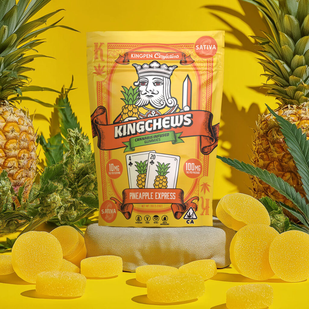 Gummy package with illustrated playing card king holding pineapple and playing cards with numbers 4 and 20 with pineapples illustrated on cards. Gummy package is surrounded by gummy tablets, real pineapples, flowering cannabis buds and leaves